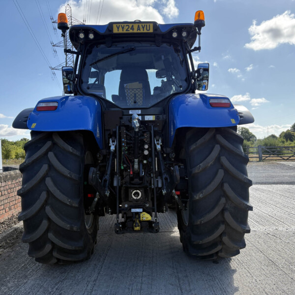 Rear view of a large blue agricultural tractor with oversized tires, hitch and hydraulic components, on a road beside a brick wall on a sunny day
