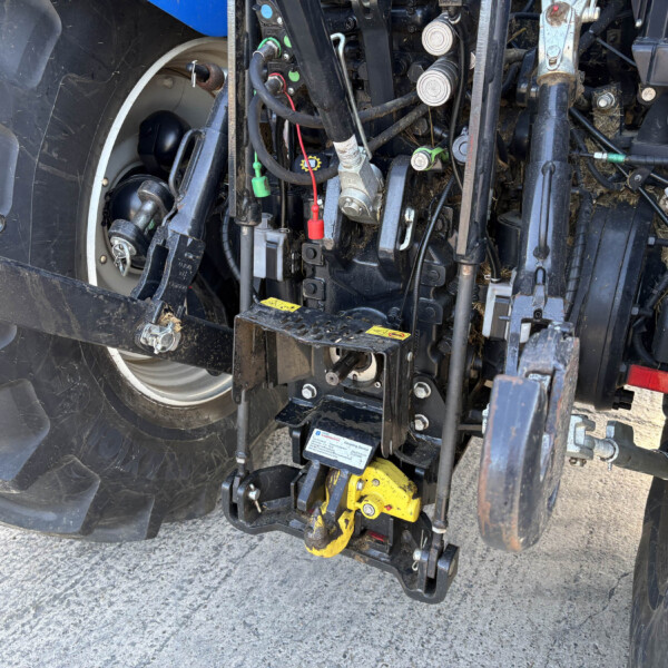 Rear tractor chassis showing hydraulic valves, hoses, and a yellow drawbar hitch with the wheel nearby