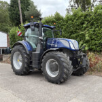 Blue New Holland tractor parked beside a hedge and a brick building with a Russell's sign in the background.