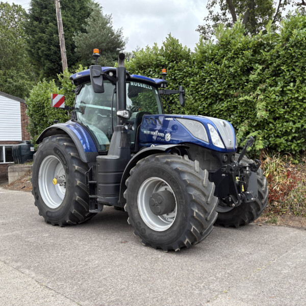 Blue New Holland tractor parked beside a hedge and a brick building with a Russell's sign in the background.