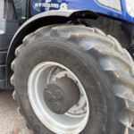 Close-up of a large tractor rear wheel with thick tread, showing dirt on the tire and a blue tractor body labeled New Holland