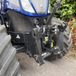 Blue tractor front with dual round headlights and hydraulic hitch exposed, large rear tires nearby in a garden setting.