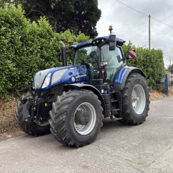 Blue New Holland tractor with large tires parked along a hedge-lined street, emergency beacons on top, brick houses in the background