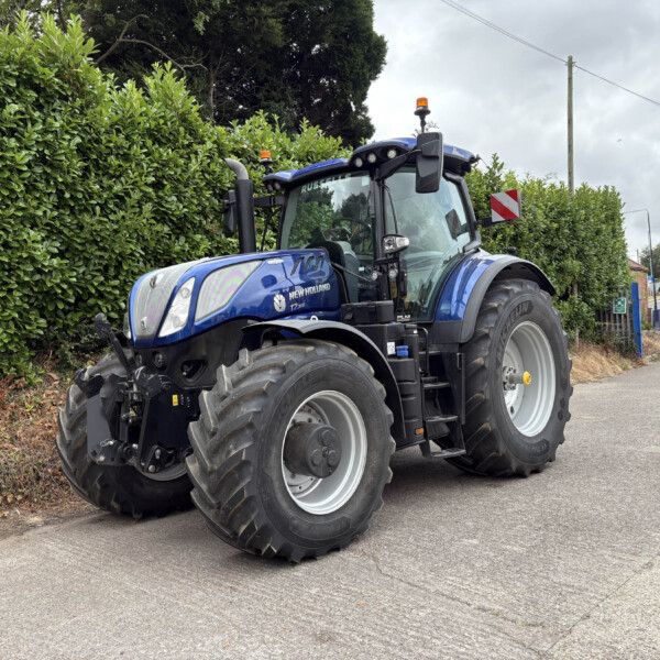 Blue New Holland tractor with large tires parked along a hedge-lined street, emergency beacons on top, brick houses in the background