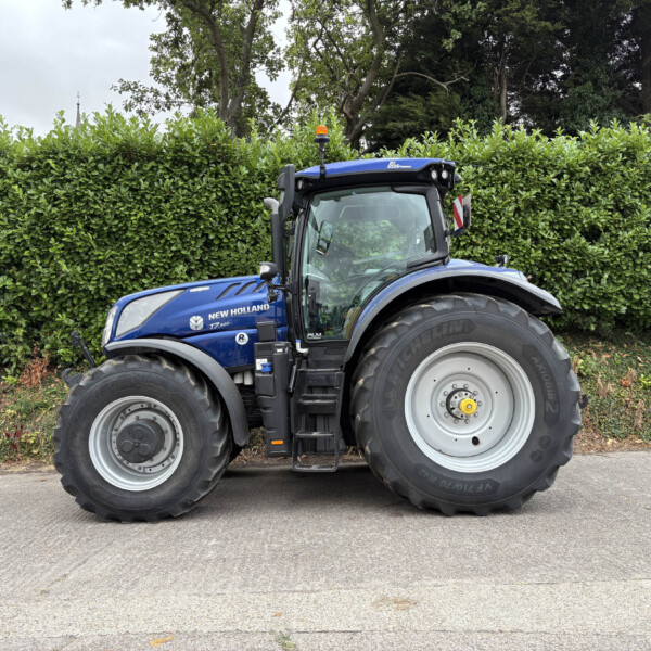 Blue New Holland tractor parked on pavement with a tall green hedge behind it, facing slightly to the left.