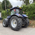 Blue New Holland tractor parked in a lot, large front tire close-up with hedges and a building in the background