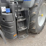 Close-up of a large truck's rear step area and wheel, showing dirty black plastic housing, a blue cap, and metal steps beside the tire rim with a yellow hub accessory nearby.