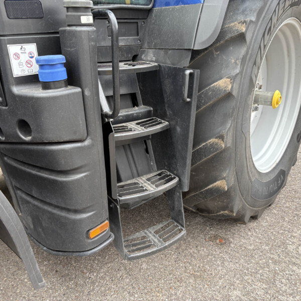 Close-up of a large truck's rear step area and wheel, showing dirty black plastic housing, a blue cap, and metal steps beside the tire rim with a yellow hub accessory nearby.