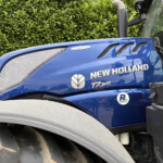 Blue New Holland T7.300 tractor side view with hood vents beside a green hedge.