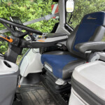 Interior of a vehicle cabin with a blue and gray driver's seat, steering wheel, and control console featuring a tablet mounted on a arm. Green hedge visible outside the windshield.