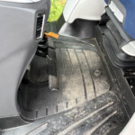 Driver's footwell showing two pedals (brake and accelerator) and a dusty rubber floor mat with an orange strap or tether on the floor nearby.