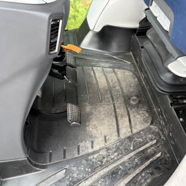 Driver's footwell showing two pedals (brake and accelerator) and a dusty rubber floor mat with an orange strap or tether on the floor nearby.