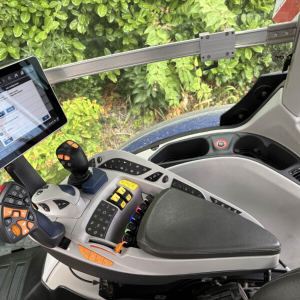 Cabin of heavy equipment with a tablet on a mount, joystick, and colorful control levers; green hedge visible through the window