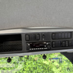 Close-up of a car dashboard center console with multiple switches and a radio, seen from the passenger seat, greenery outside the windshield.