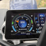 Close-up of a vehicle's digital instrument cluster showing a large circular speed/RPM gauge, blue accent markings, and fuel indicators with BLUEPOWER branding on the center panel.