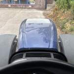 Driver's-eye view from inside a blue vehicle looking over the hood at a brick building and greenery outside.