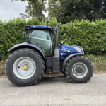 Blue New Holland tractor parked on a paved road beside a tall green hedge, front wheel and cabin visible.