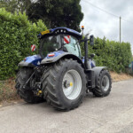 Blue tractor parked along a hedge on a residential street with overcast sky in the background.