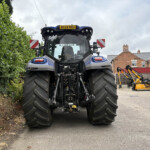Rear view of a blue tractor parked in a lot, with a tall hedge on the left and orange farming equipment on the right.