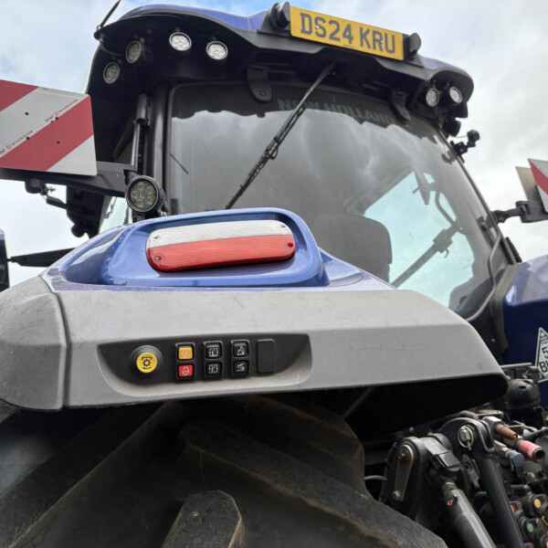Close-up of the front of a blue New Holland tractor, showing the dashboard controls, red light, and a yellow license plate DS24 KRU against a cloudy sky.