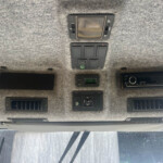 Overhead vehicle ceiling with multiple switches, two square lights, and a stereo head unit mounted below in a gray fabric panel.