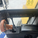 Inside the cabin of a construction vehicle, showing a control panel with many labeled buttons and an orange joystick nearby