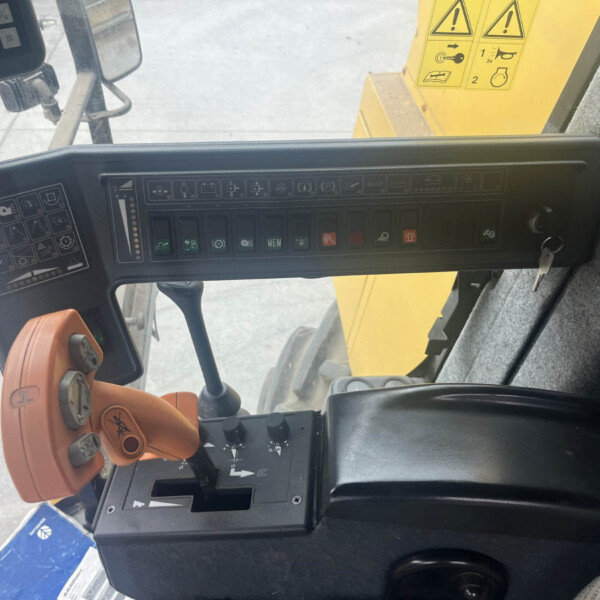 Inside the cabin of a construction vehicle, showing a control panel with many labeled buttons and an orange joystick nearby