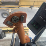 Close-up of an orange construction joystick with several grey buttons inside a cab, a yellow crane arm visible outside the windshield.