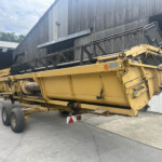 Yellow agricultural machine trailer parked beside a corrugated metal warehouse, large conveyor header visible in foreground.