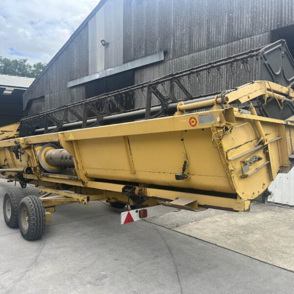 Yellow agricultural machine trailer parked beside a corrugated metal warehouse, large conveyor header visible in foreground.