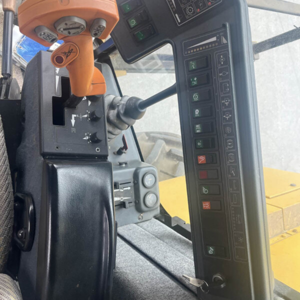 Orange hydraulic joystick and control panel inside heavy equipment cab with numerous labeled switches and gauges.