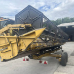 Yellow heavy construction machinery parked in an outdoor yard, with a long black conveyor-like arm extended over it and a covered building in the background.