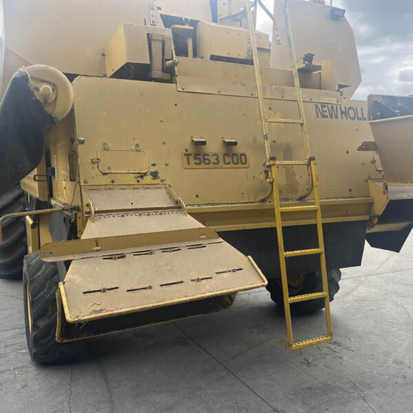 Rear view of a large yellow New Holland industrial machine with a metal ladder and chute in an outdoor yard.