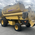 Side view of a yellow New Holland TF78 combine harvester parked in an industrial yard with large tires and ladder.