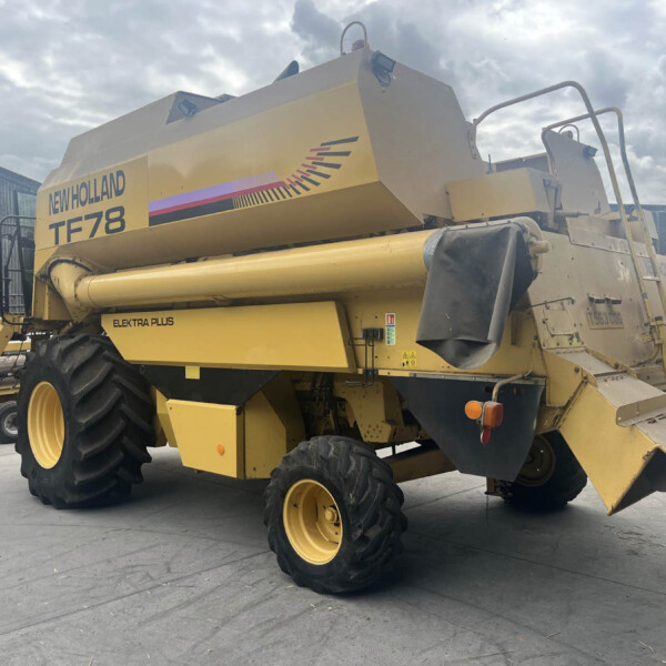 Side view of a yellow New Holland TF78 combine harvester parked in an industrial yard with large tires and ladder.