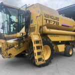 Front view of a large yellow New Holland TF78 combine harvester with an open cab, ladder, and oversized tires parked outdoors near a shed.