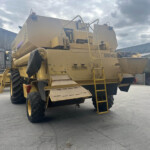 Large yellow New Holland combine harvester with ladder and platforms, parked in an outdoor industrial yard near corrugated metal buildings.
