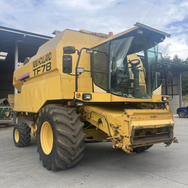 Side/front view of a yellow New Holland TF78 combine harvester parked outdoors, showing large tires and cabin.