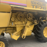 Close-up of the belt-driven gear train on a yellow New Holland harvest machine, showing exposed pulleys and large wheels in the frame.