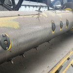 Close-up of a large, dusty gray industrial pipe with yellow patches, bolts and brackets, and frayed wire resting on its surface on a metal platform.
