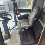 Interior of a heavy construction vehicle cab with steering wheel, orange joystick, and row of control switches; driver seat to the right and warning stickers on the window visible outside.