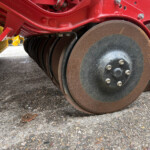 Close-up of large rusty steel rollers beneath a red agricultural implement on a gravel surface.