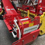 Close-up of a red Pöttinger Lion 3002 agricultural implement with yellow gears and multiple warning decals visible on the frame.