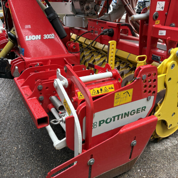 Close-up of a red Pöttinger Lion 3002 agricultural implement with yellow gears and multiple warning decals visible on the frame.