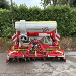 Front view of a Pöttinger seed drill mounted on a red frame, parked on a paved surface with a dense green hedge behind it.
