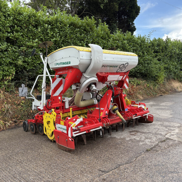 Pöttinger seed drill with white hopper and red frame parked beside a tall green hedge on a road.