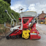 Red and white Pöttinger agricultural implement with yellow wheels, parked beside a green hedge and brick houses in the background.