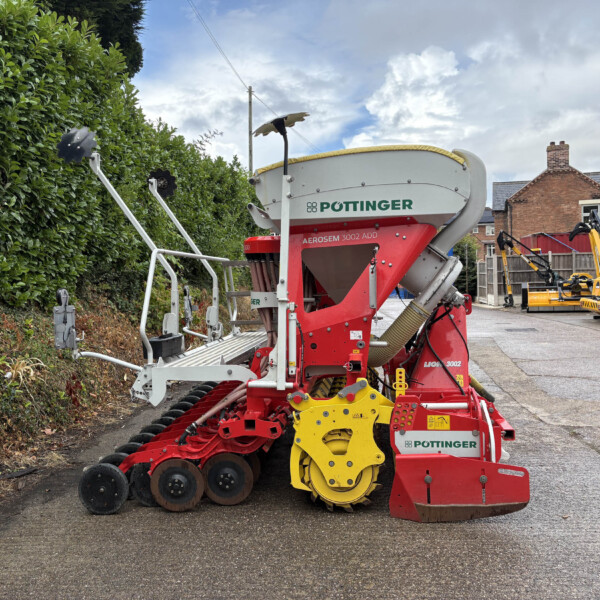 Red and white Pöttinger agricultural implement with yellow wheels, parked beside a green hedge and brick houses in the background.
