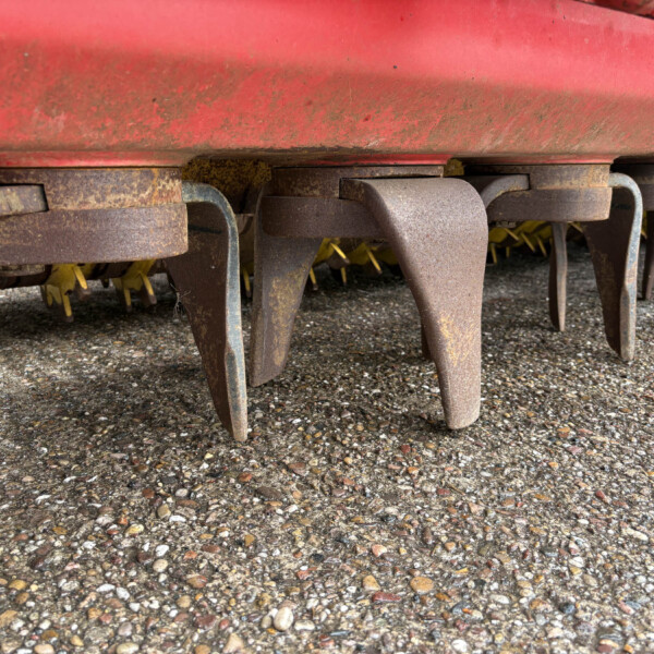 Close-up of rusted metal tines or prongs under a red farming implement resting on a pebbled ground.