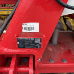 Red metal frame of agricultural equipment with a barcode label, a black Pöttinger plate, and hanging chains on the right side. Water droplets dot the surface, with yellow machinery parts faintly in the background.